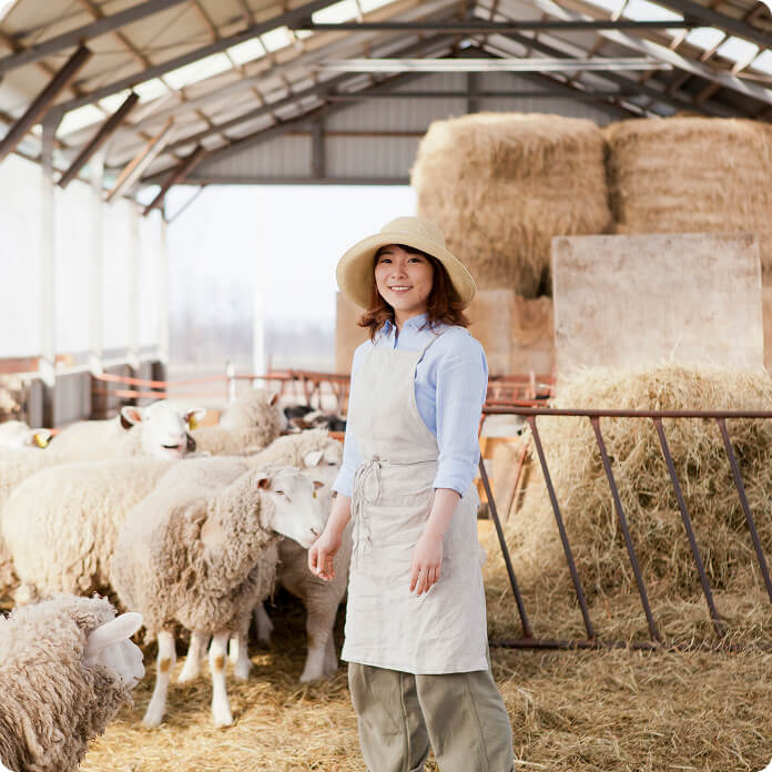 羊と牧草をバックにエプロン姿で立つ笑顔の女性の写真
