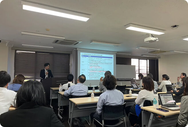 会議室のような場所でスクリーンを前に話している人とそれを聞く人たちの写真